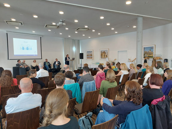 Ein großer Raum mit ca. 50 Menschen. Es ist eine Präsentation an der Wand und eine Gesprächsrunde auf einem Podium aus 4 Gästen.