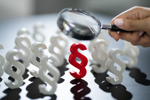 Mit einer Lupe wird ein rotes Paragrafenzeichen untersucht.